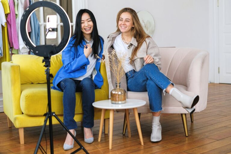 Two stylish women seated on a sofa, vlogging with a smartphone and ring light in a cozy indoor setting.